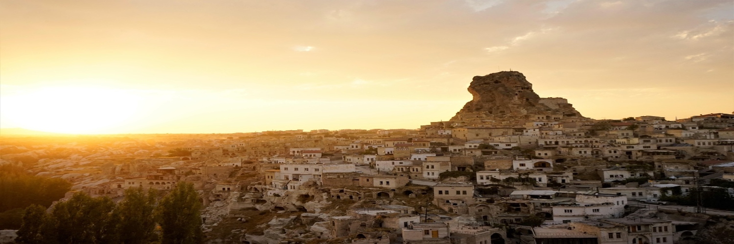 Golden sunset over Cappadocia, Turkey, with silhouettes of fairy chimneys and ancient buildings against a colorful sky.