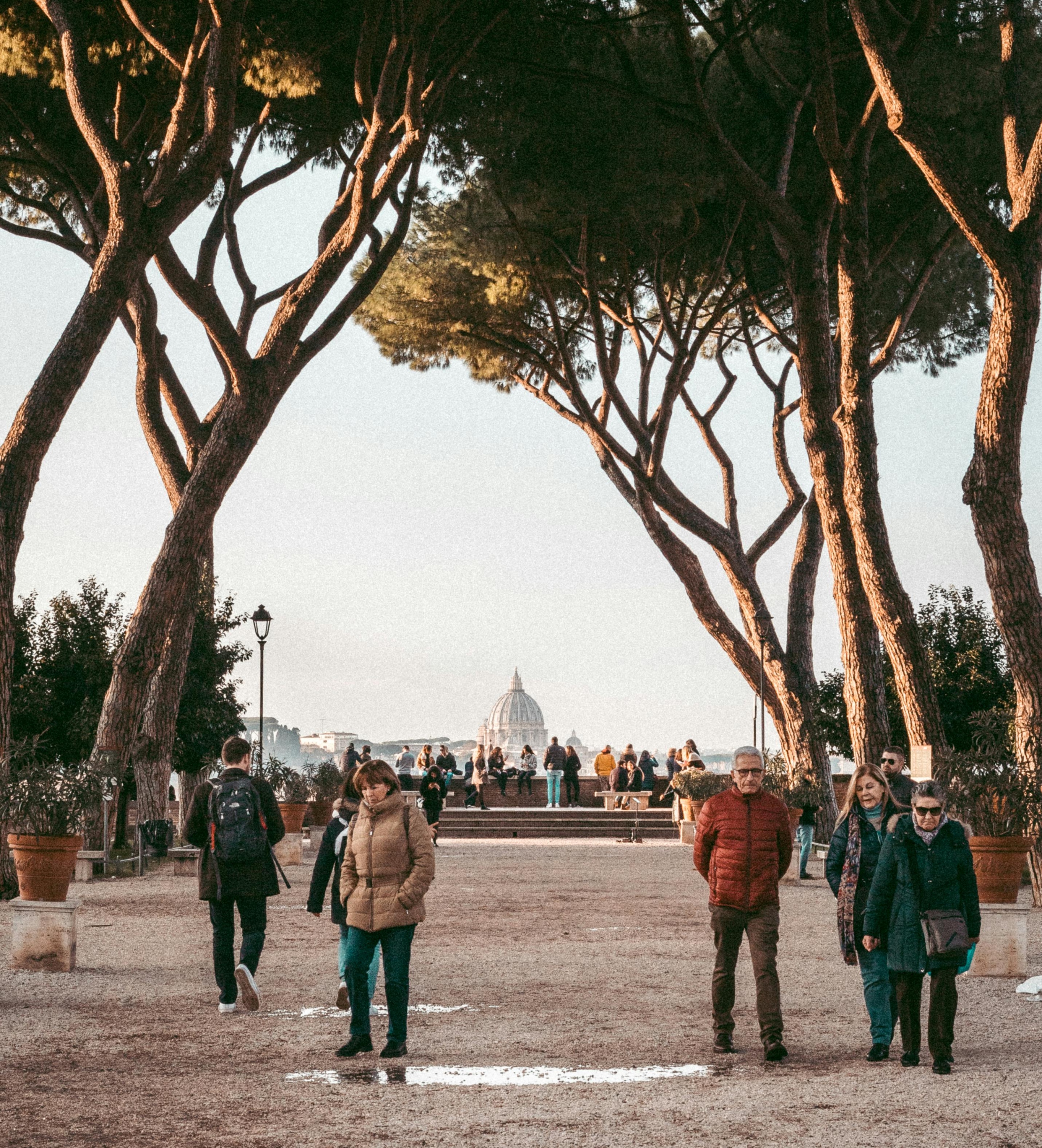 A winter scene in Rome featuring people strolling through a park, surrounded by trees and a distant building.