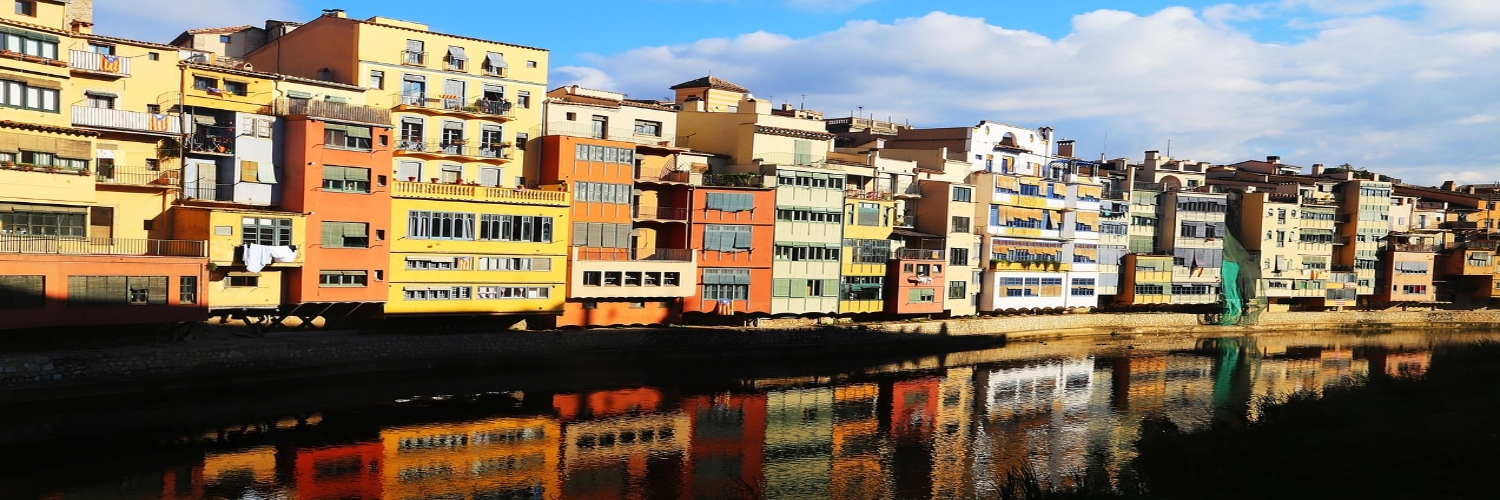 A serene body of water reflecting the skyline of Girona town, surrounded by lush greenery and historic architecture.