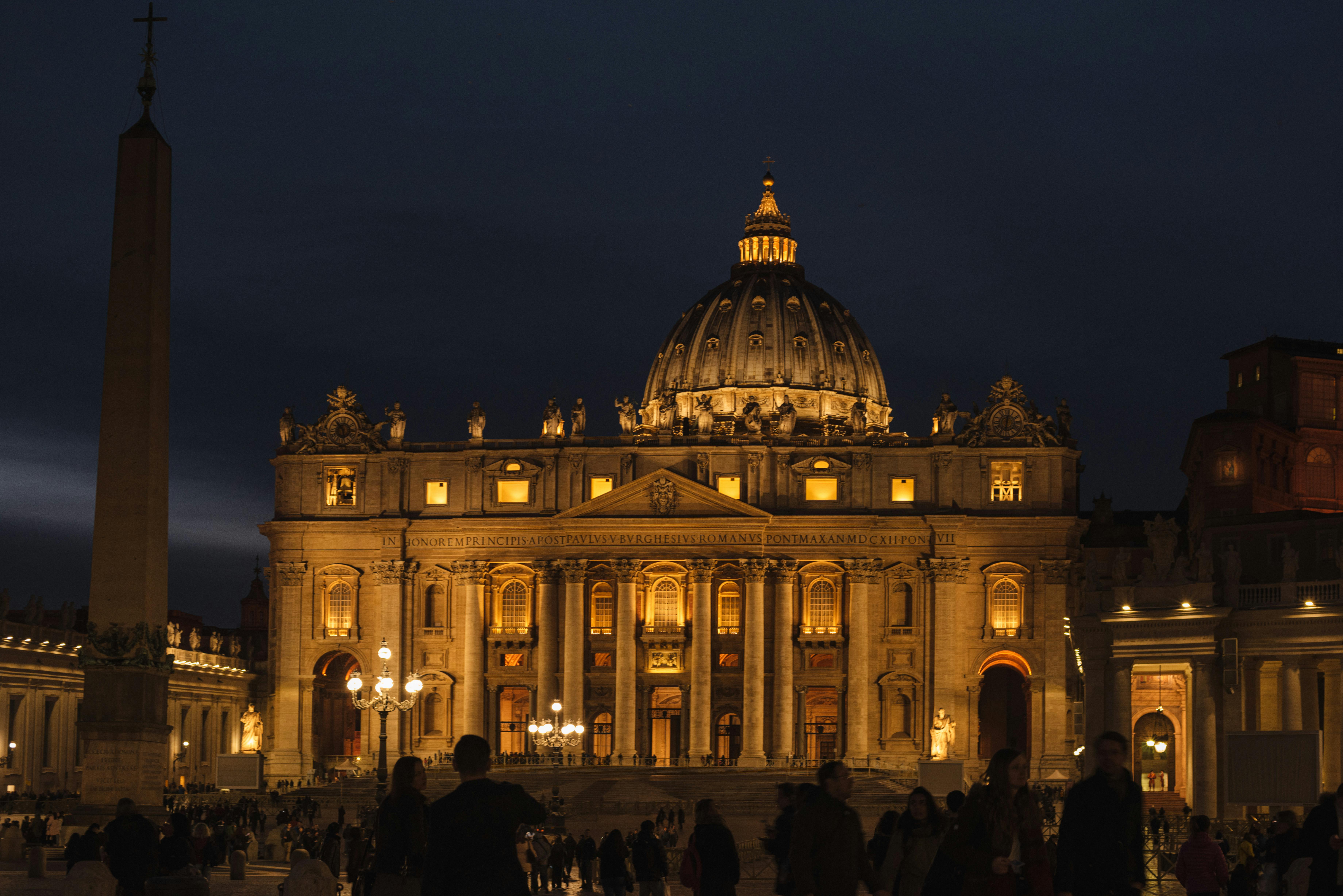Nighttime view of Saint Peter’s Basilica as people walk by in Rome.