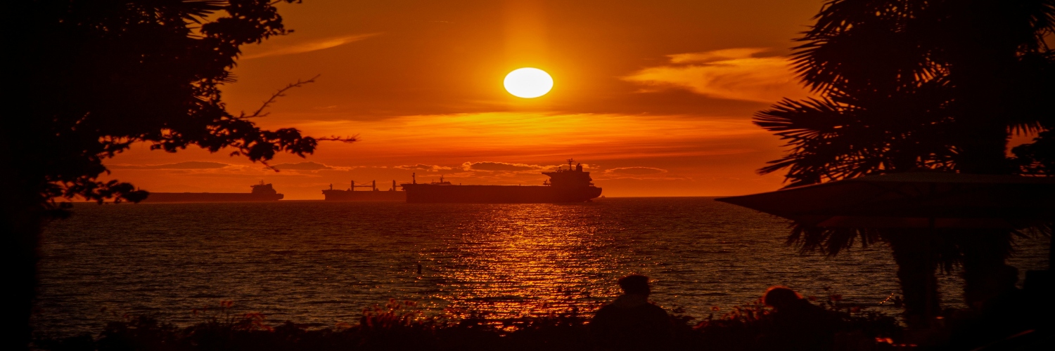 A large ship sails on the water at sunset, with the Vancouver skyline visible in the background.