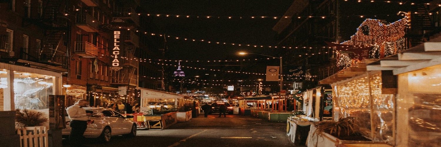 A festive New York city street at night, adorned with twinkling lights strung overhead for Christmas celebrations.