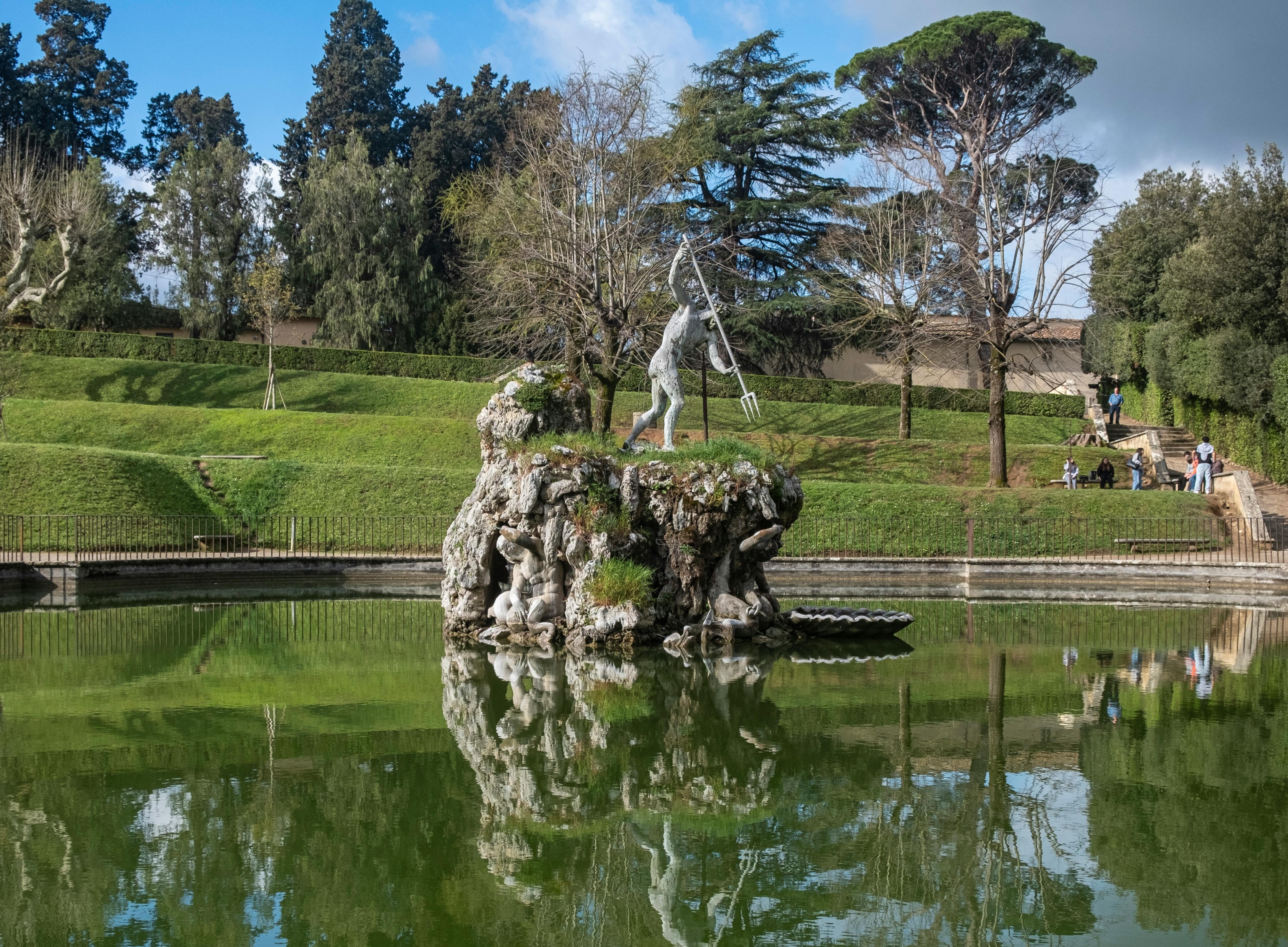 A statue of a man stands in the center of a pond at the Boboli Gardens in Florence, surrounded by lush greenery.
