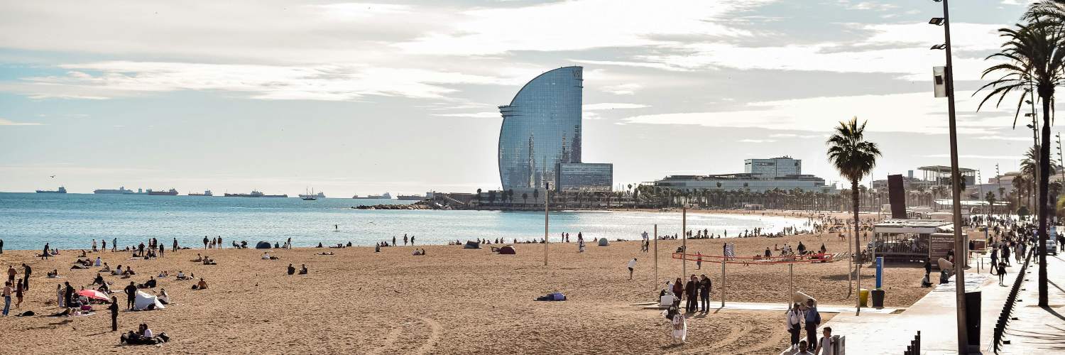 A sunny day at Barceloneta Beach, with people walking along the shore and a tall building in the background.