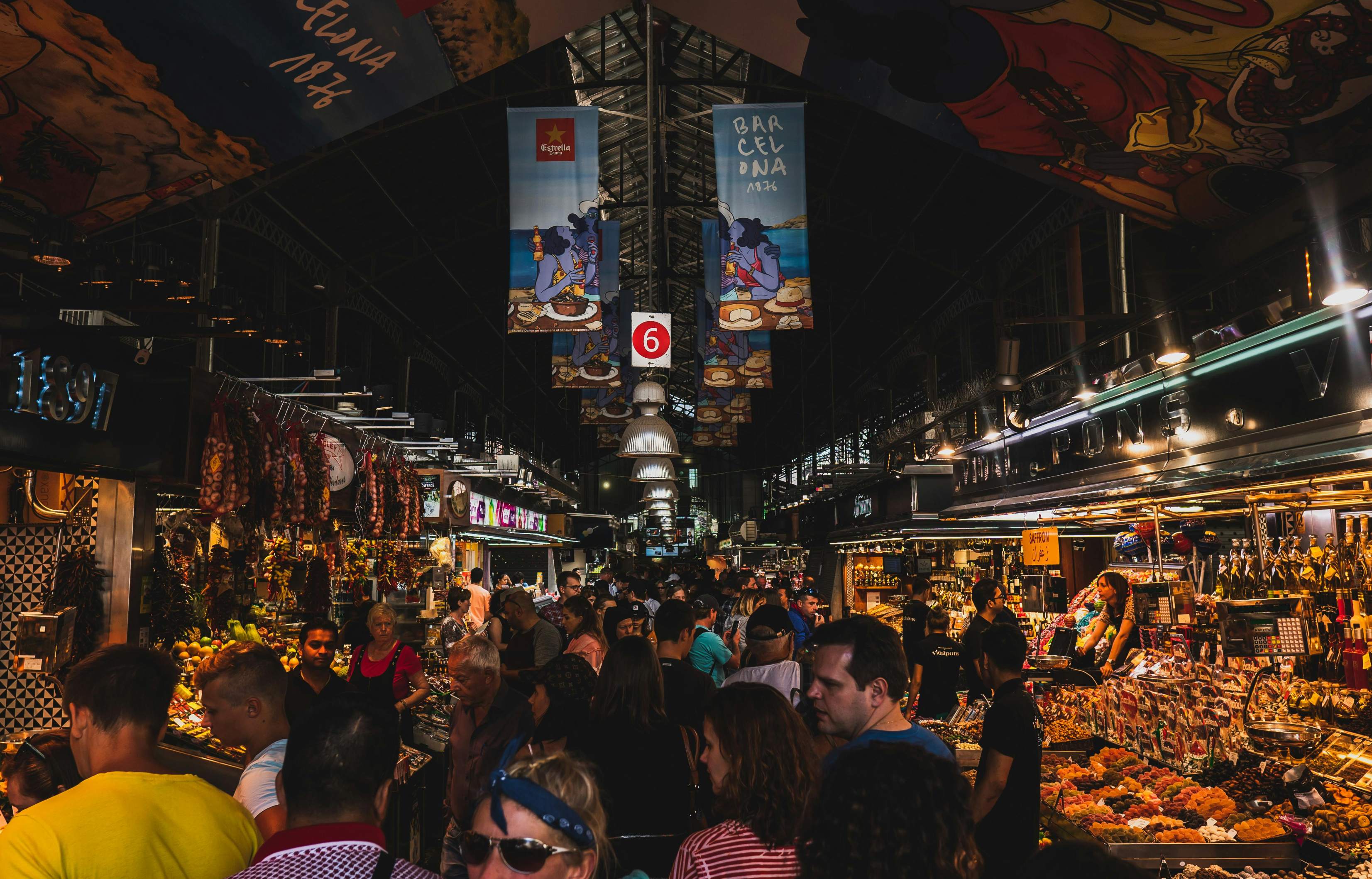 A bustling Barcelona market filled with people walking among colorful stalls and vibrant displays of goods.
