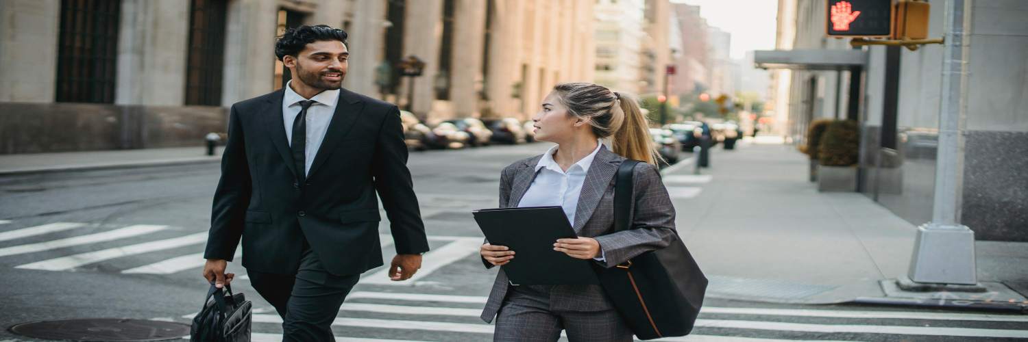 Two business travelers walking down a city street, dressed in professional attire, engaged in conversation.