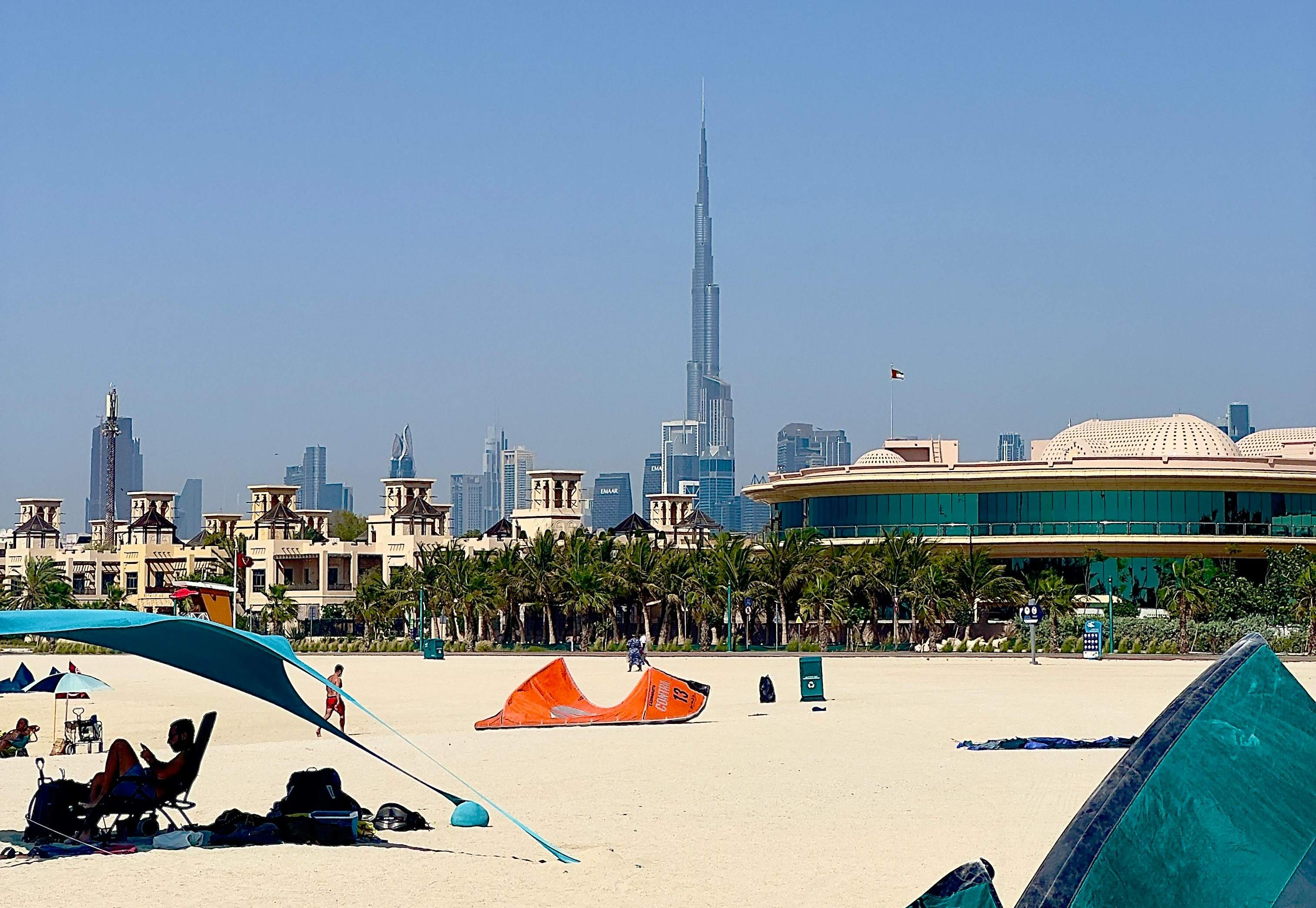 A beachfront view in Dubai featuring tents, people on the sand, and the Burj Khalifa in the background.
