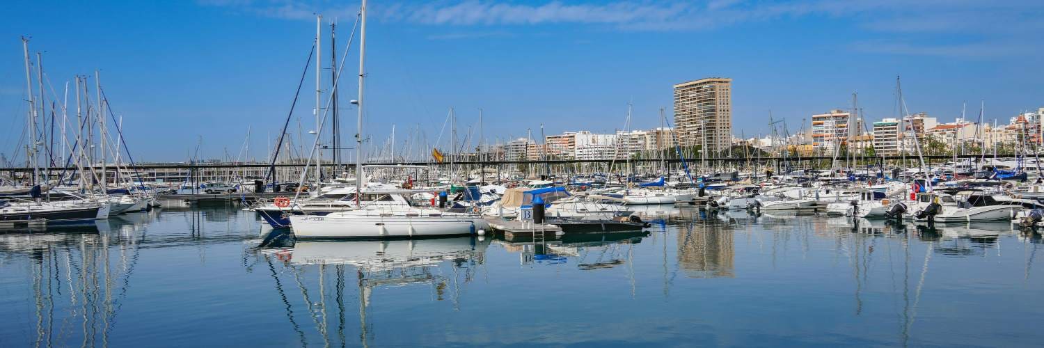Boats and yachts docked at Alicante marina with calm water reflections and city buildings under a clear blue sky.
