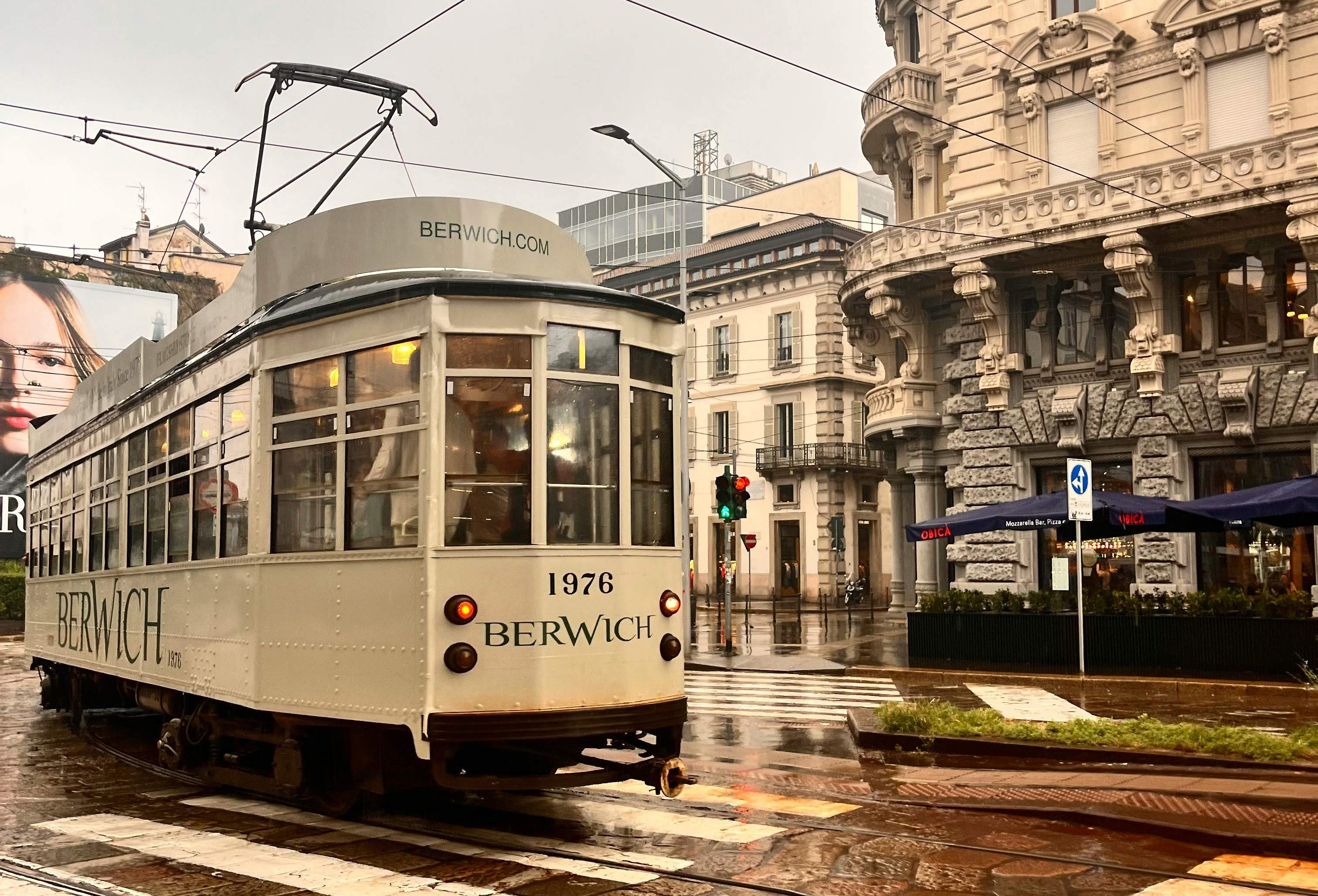 A vintage white trolley car travels along a rainy street in Milan, surrounded by wet pavement and blurred city scenery.
