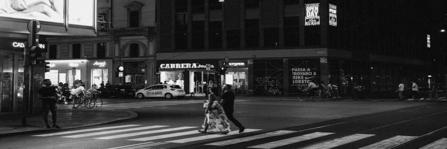 A couple walking hand in hand across a bustling street in downtown Milan at night, with bright lights and cars nearby.