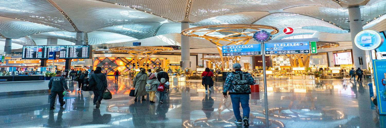 People walking through a busy airport terminal, surrounded by various signs, indicating transfer costs and directions.