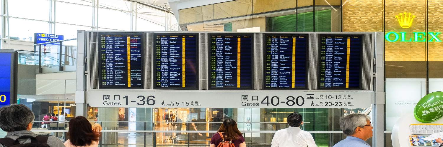 Travelers with luggage walk through an airport, passing a large screen displaying flight tracking information.