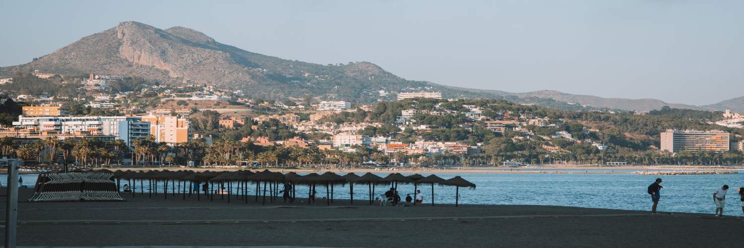 People stroll along Málaga Beach, with gentle waves lapping at the shore and mountains in the background.