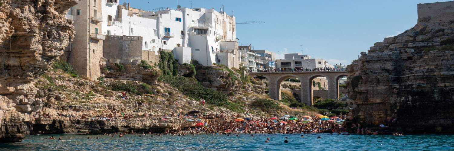 In Polignano, Italy, individuals swim in the water beside a dramatic cliff, capturing the beauty of the coastline.