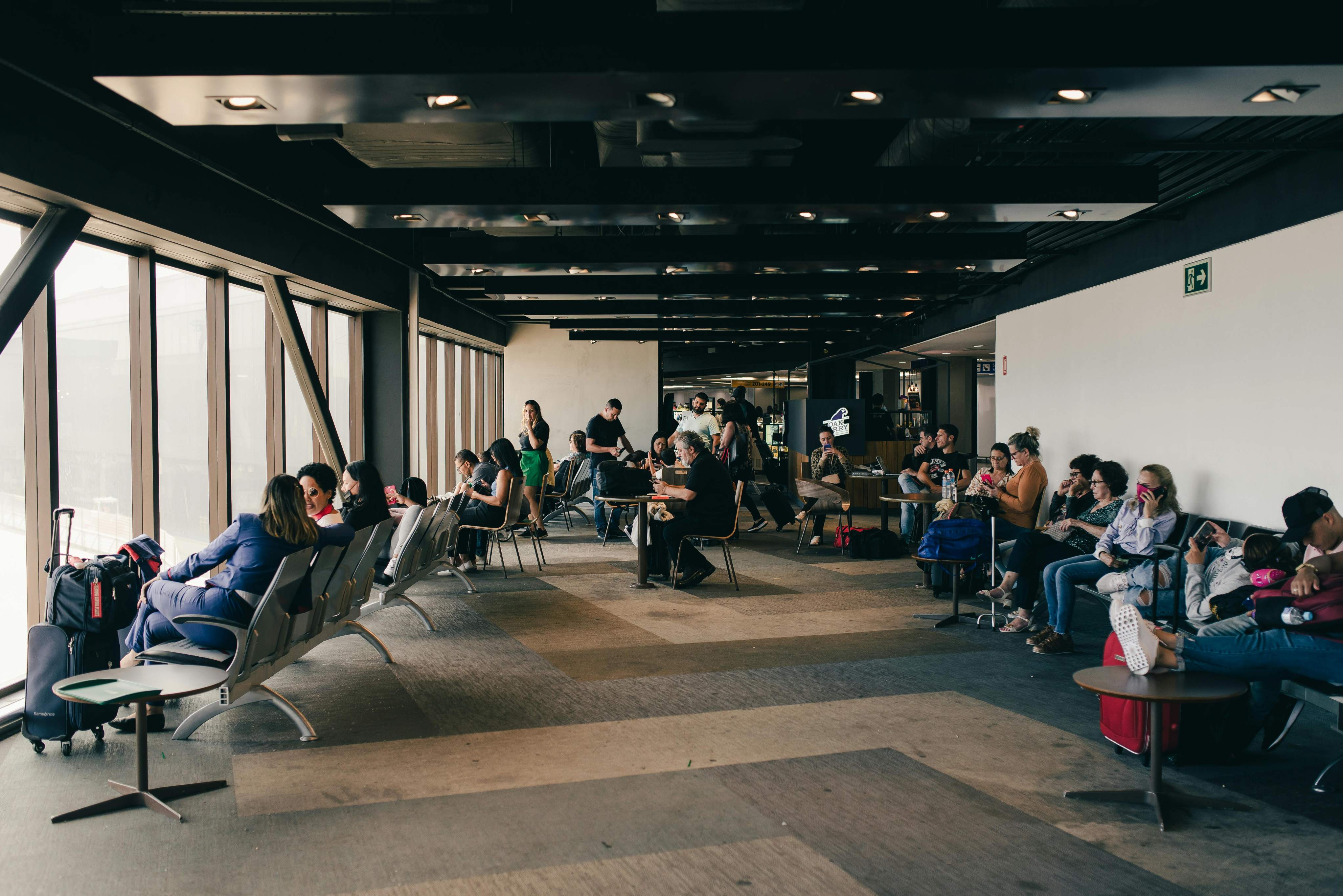 Travelers in an airport waiting area, seated and waiting, with luggage nearby and screens displaying flight information.