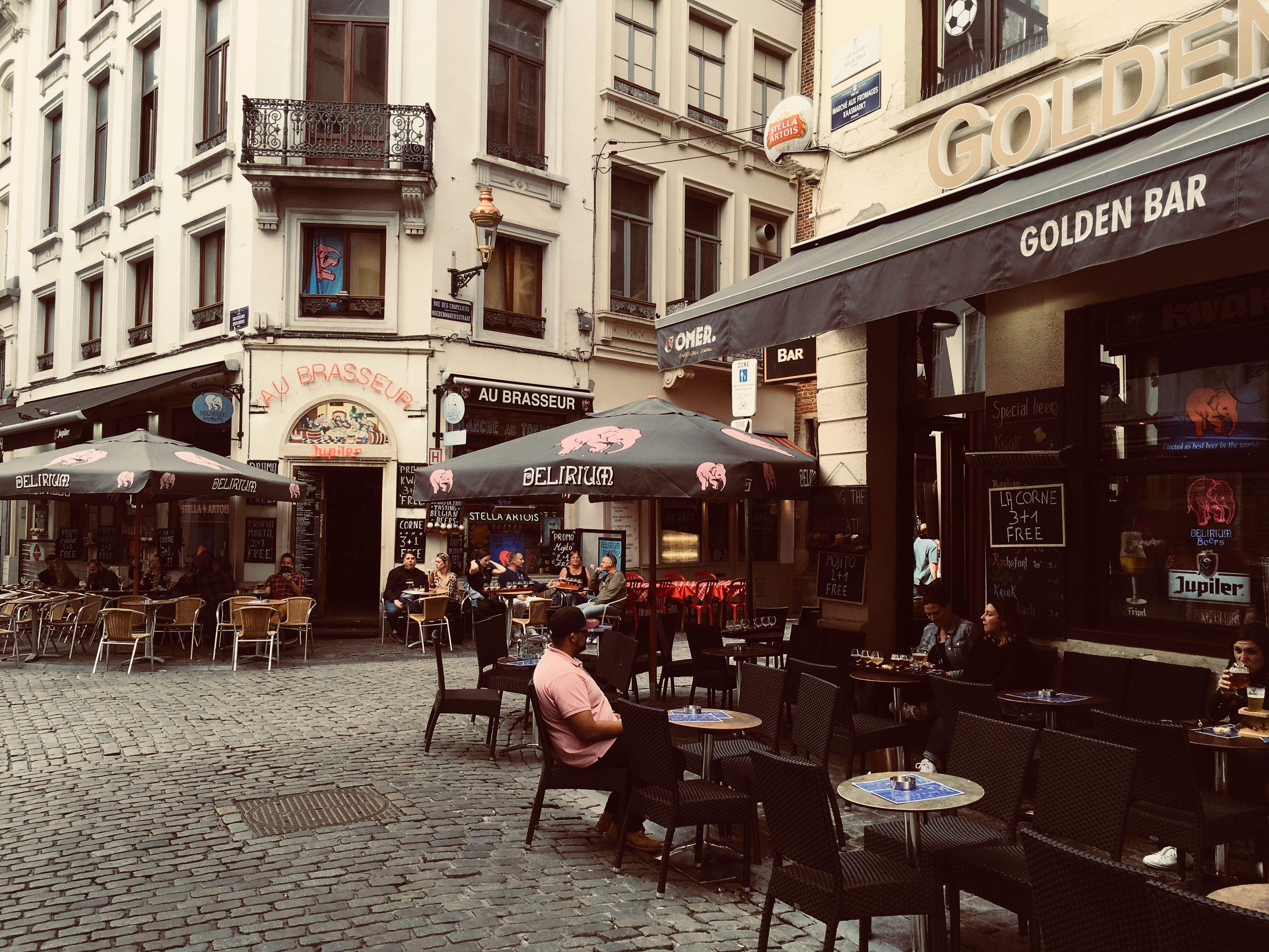 Outdoor cafes line a brick-paved street in Brussels, with patrons enjoying drinks and meals under colorful umbrellas.