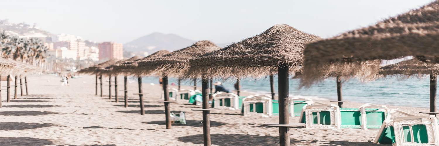 Sandy beach in Malaga with straw sun umbrellas and seaside lounge chairs under a clear sky.