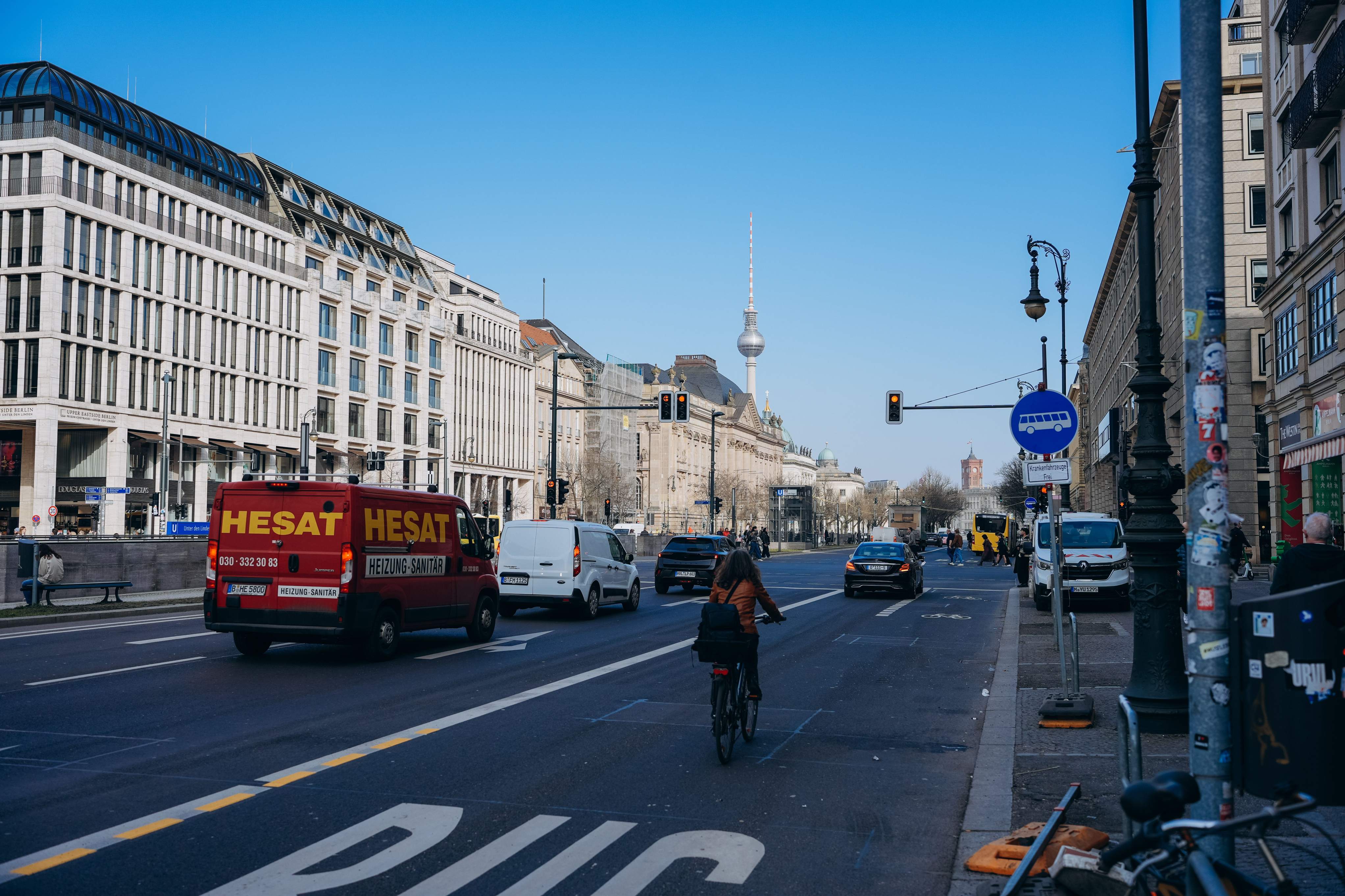 A woman riding a bicycle along a busy street in Berlin, surrounded by urban buildings and pedestrians.