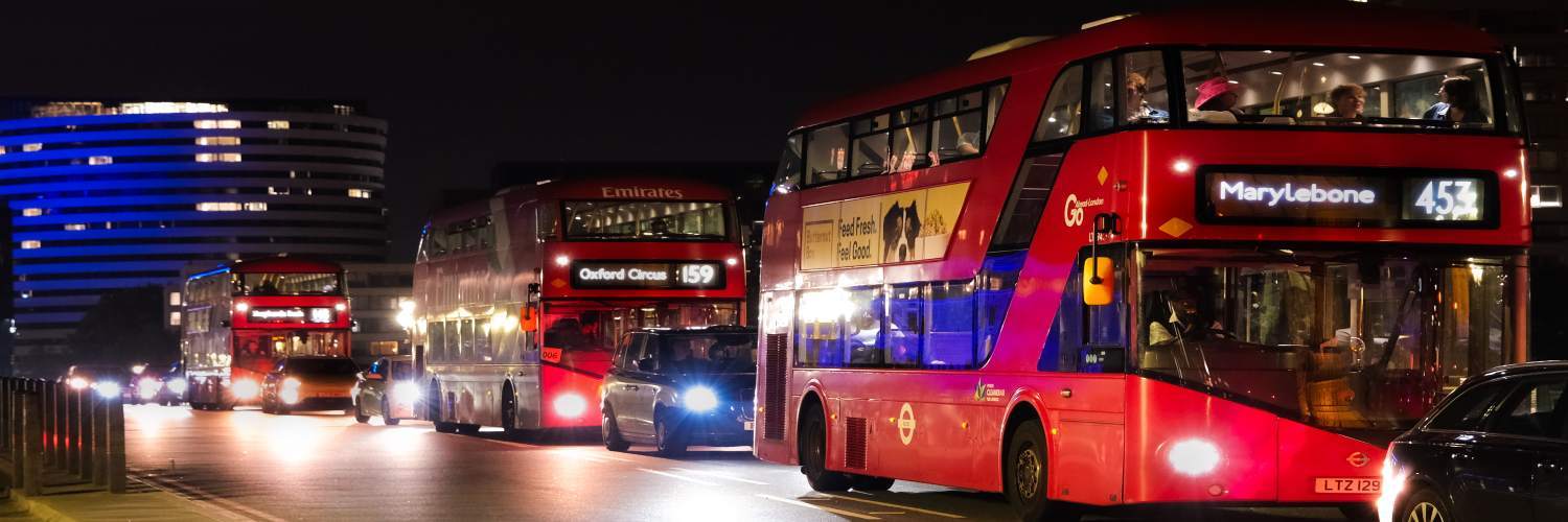 A red double-decker bus travels down a city street at night, illuminated by streetlights and surrounding buildings.