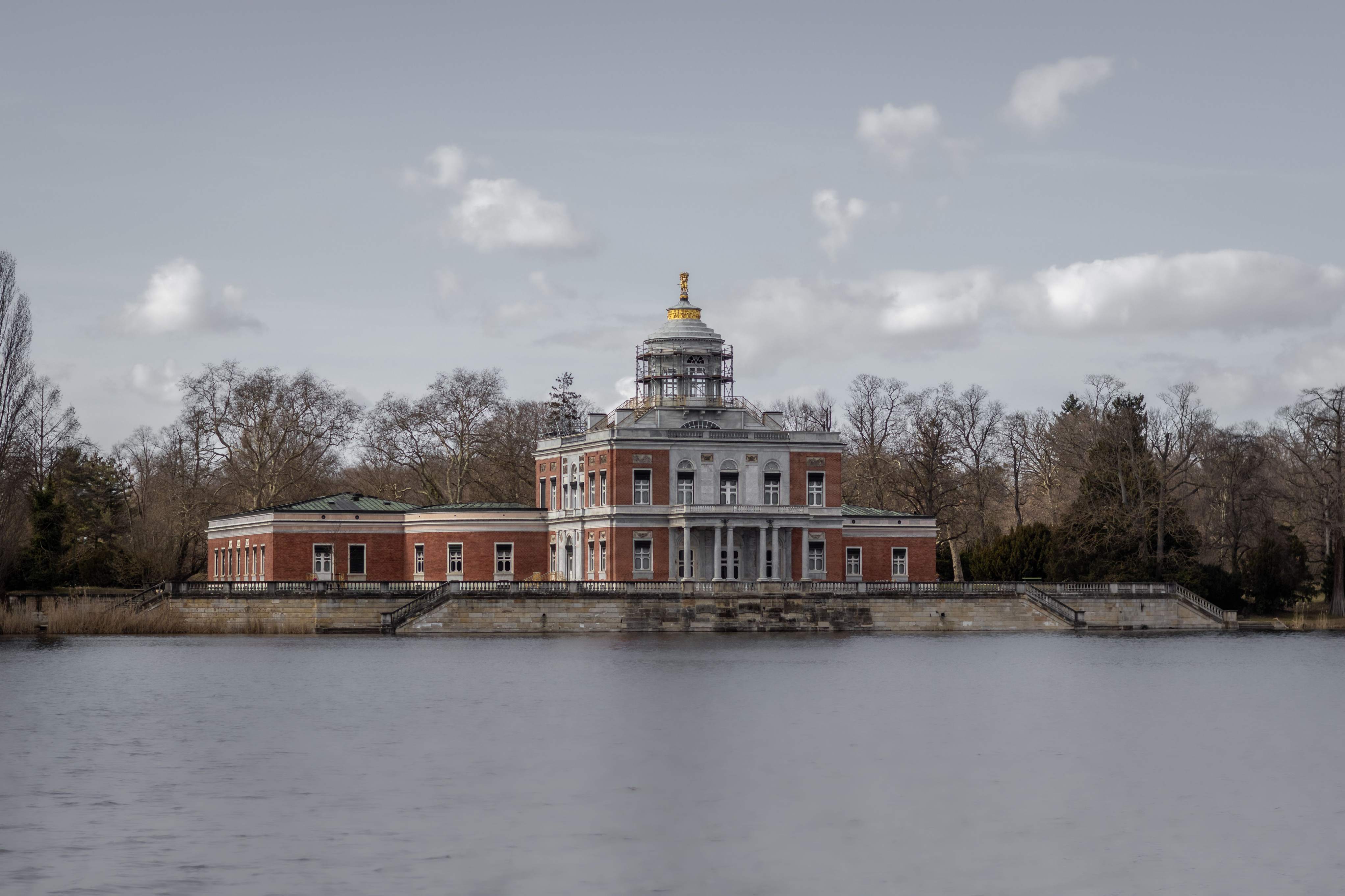A serene lake reflecting the Palace of Hesse in Potsdam, surrounded by lush greenery and a clear blue sky.