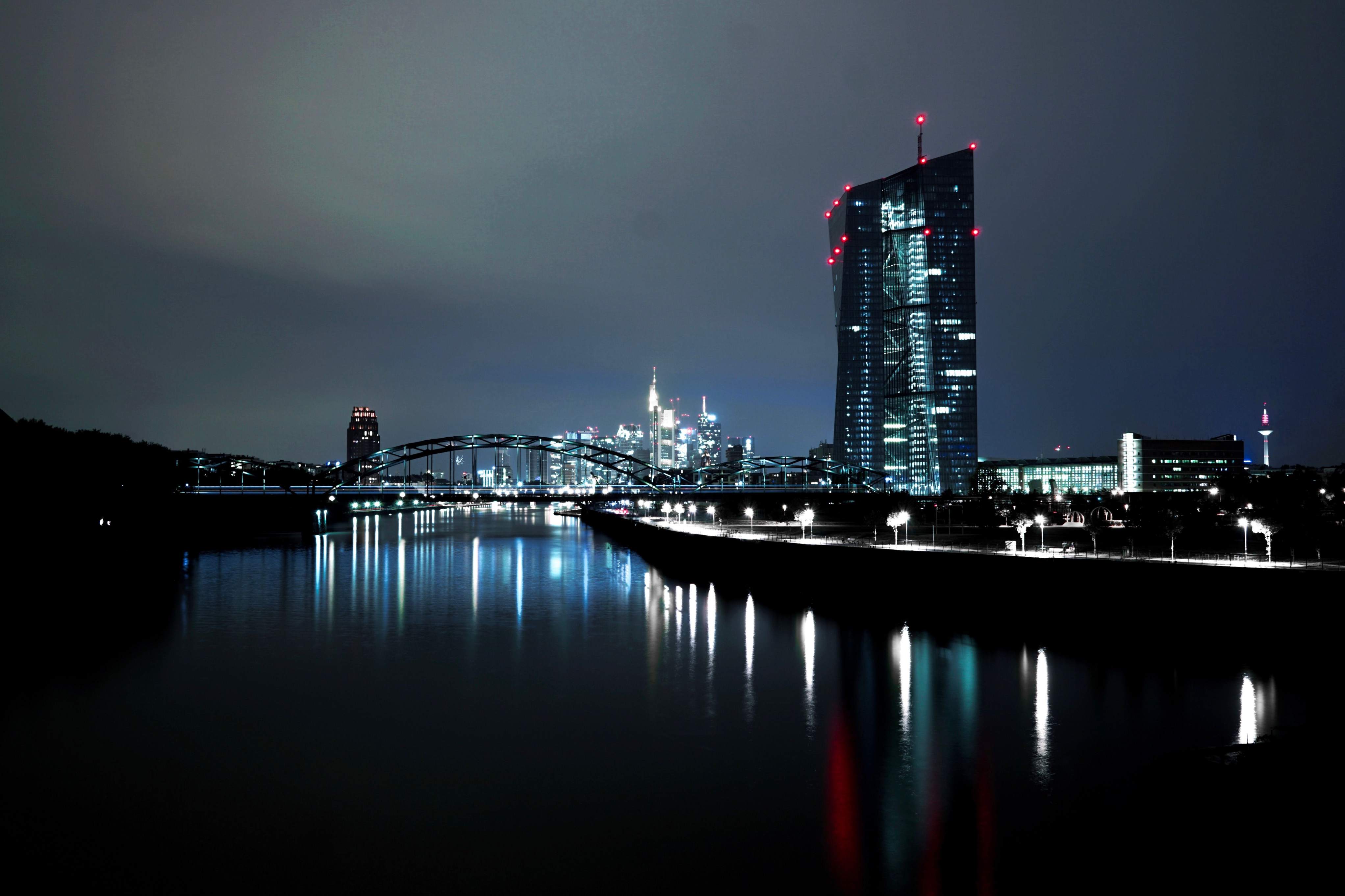Frankfurt's skyline at night, featuring illuminated skyscrapers, a river, and a bridge reflecting city lights.