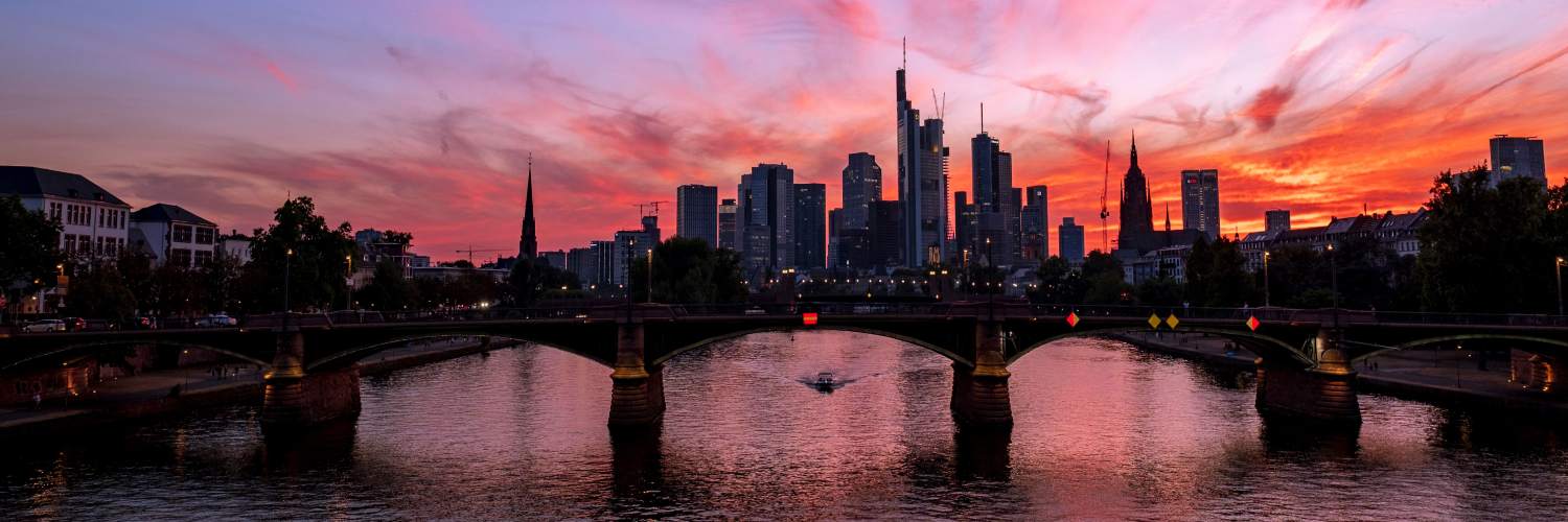 Sunset over Frankfurt's skyline, showcasing tall buildings silhouetted against a vibrant orange and purple sky.