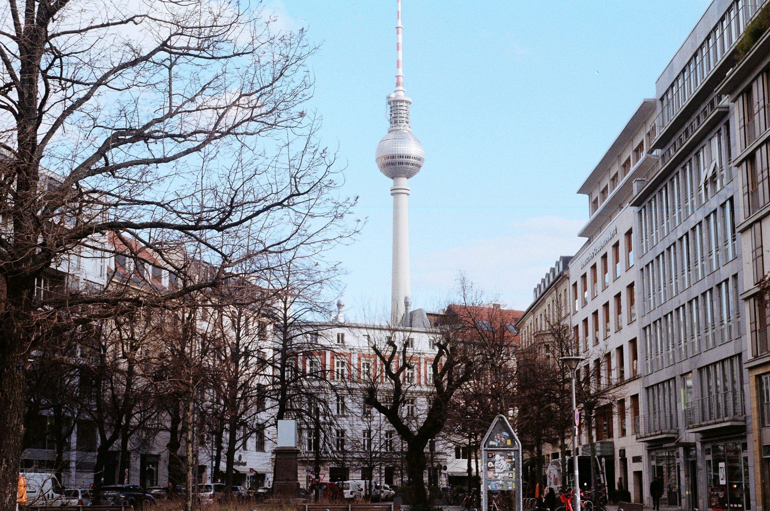 A city street scene featuring the Berlin TV Tower prominently in the background against a clear sky.