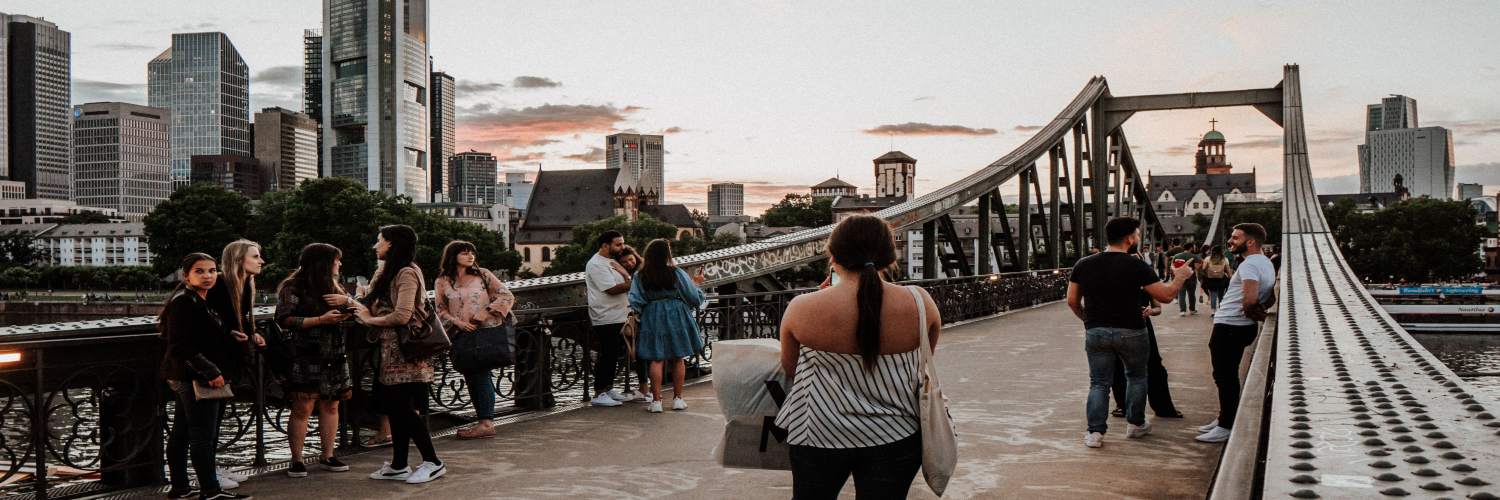 People walking on Eiserner Bridge in Frankfurt, with city buildings and a river visible in the background.