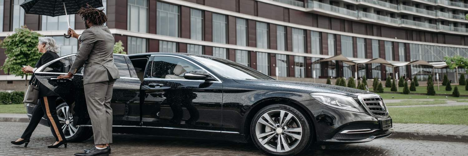 A businesswoman in formal attire holds an umbrella over a luxury car, indicating a chauffeur service in Berlin.